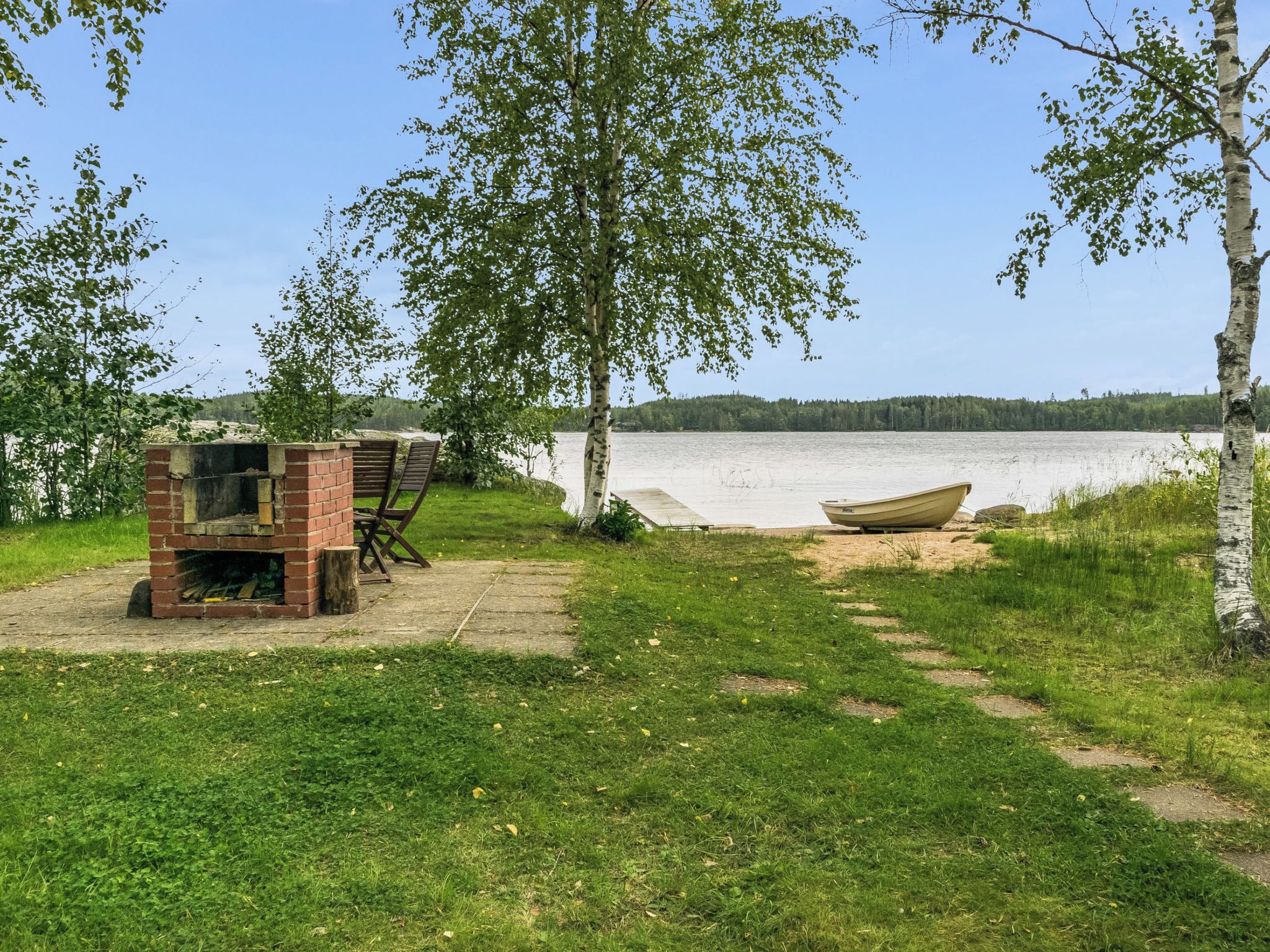 Een landschap aan het meer met een grill van baksteen en houten stoelen, een boot aan de oever en een helder uitzicht over het water.