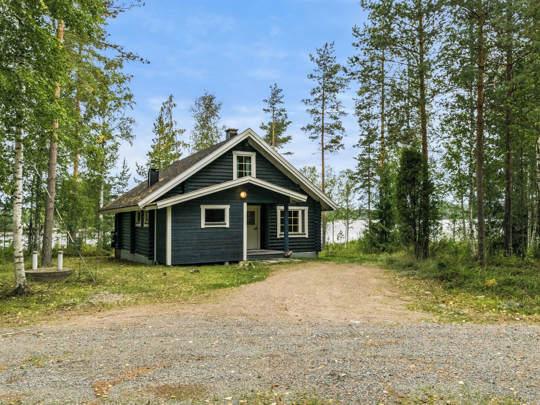 Mökki, jonka edessä on sorapäällysteinen ajotie, ja jonka ympärillä kasvaa puita.