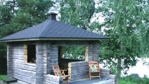 A rustic stone cabin with a sloped roof, a porch with two wooden chairs, and a painting leaning against the wall. - Rantamökki by Interhome (Luhanka)