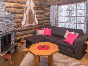 L'intérieur d'une cabane en rondins, avec un canapé d'angle, une cheminée et une grande fenêtre donnant sur des arbres couverts de neige.
