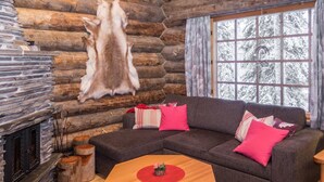 L’intérieur d’un chalet confortable, avec des murs en rondins de bois, une cheminée en pierre, une grande fenêtre donnant sur un paysage enneigé, un grand canapé d’angle gris agrémenté de coussins rouges et une table basse en bois.
