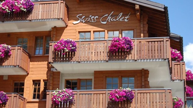 Ein hölzernes Gebäude mit Balkonen, die mit violetten Blumen geschmückt sind, und der Aufschrift "Swiss Chalet".