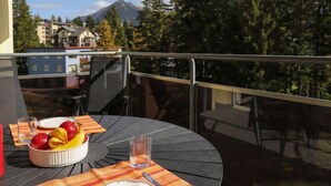 A round table with a fruit bowl, a glass of water, and a plate on a balcony overlooking a forest and buildings. - Allod Park Haus C 301 by Interhome (Davos)