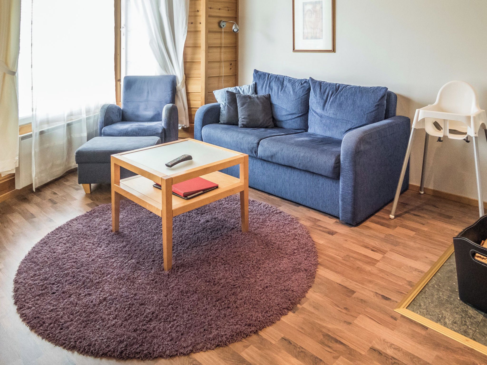 A living room with a blue sofa, a matching armchair, a small wooden coffee table with a remote control and a red book, and a purple rug.