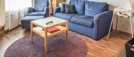 Ein Wohnzimmer mit einem blauen Sofa, einem passenden Sessel, einem kleinen Holztisch mit Fernbedienung und einem roten Buch sowie einem violetten Teppich.