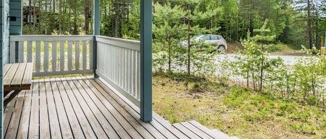 A wooden deck with a railing, overlooking a forested area with a car parked in the background.