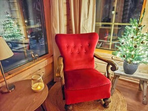 A red upholstered chair with wooden armrests, a small table with a candle, and a Christmas tree in a window.