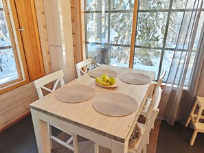 A dining area with a wooden table, white chairs, and a view of snow-covered trees through large windows.