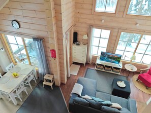 Un intérieur de chalet chaleureux avec un coin repas, un canapé, une télévision et de grandes fenêtres donnant sur le paysage enneigé.