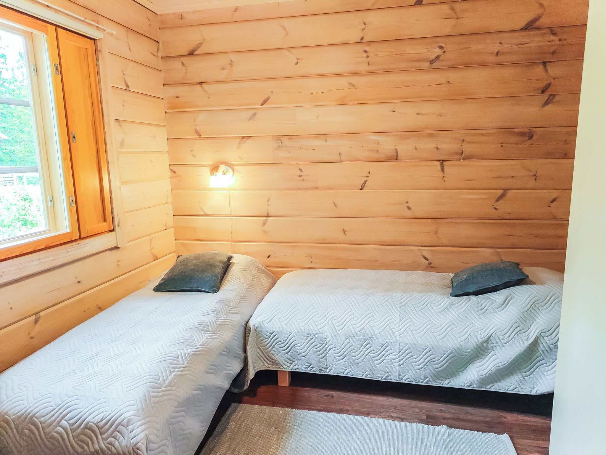 A small wooden cabin room with two beds, a window, and a wall-mounted light.