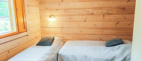 A small wooden cabin room with two beds, a window, and a wall-mounted light.