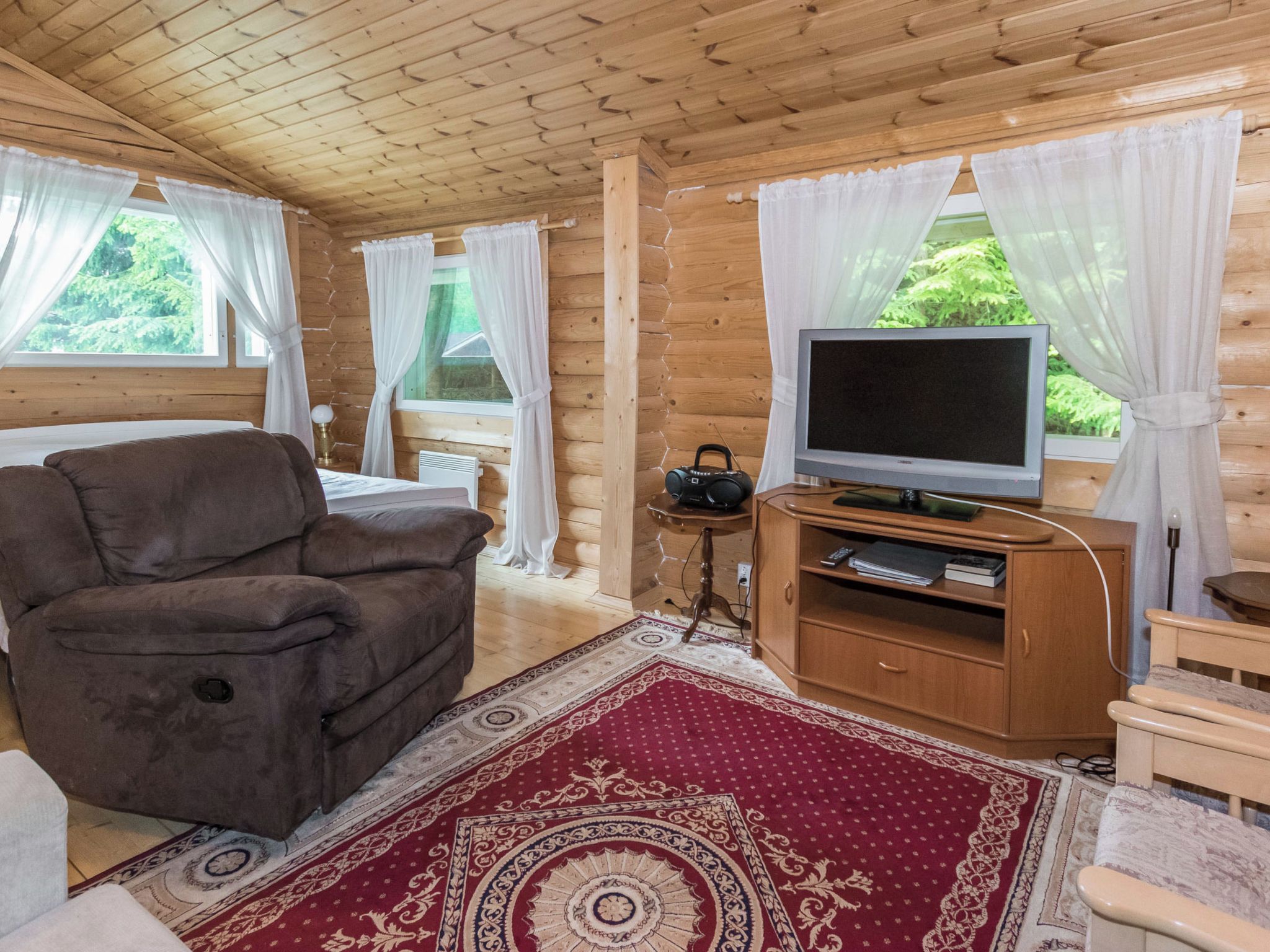 Une pièce chaleureuse dans un chalet en rondins, avec un canapé marron, un téléviseur posé sur un meuble en bois et un tapis rouge à motifs.