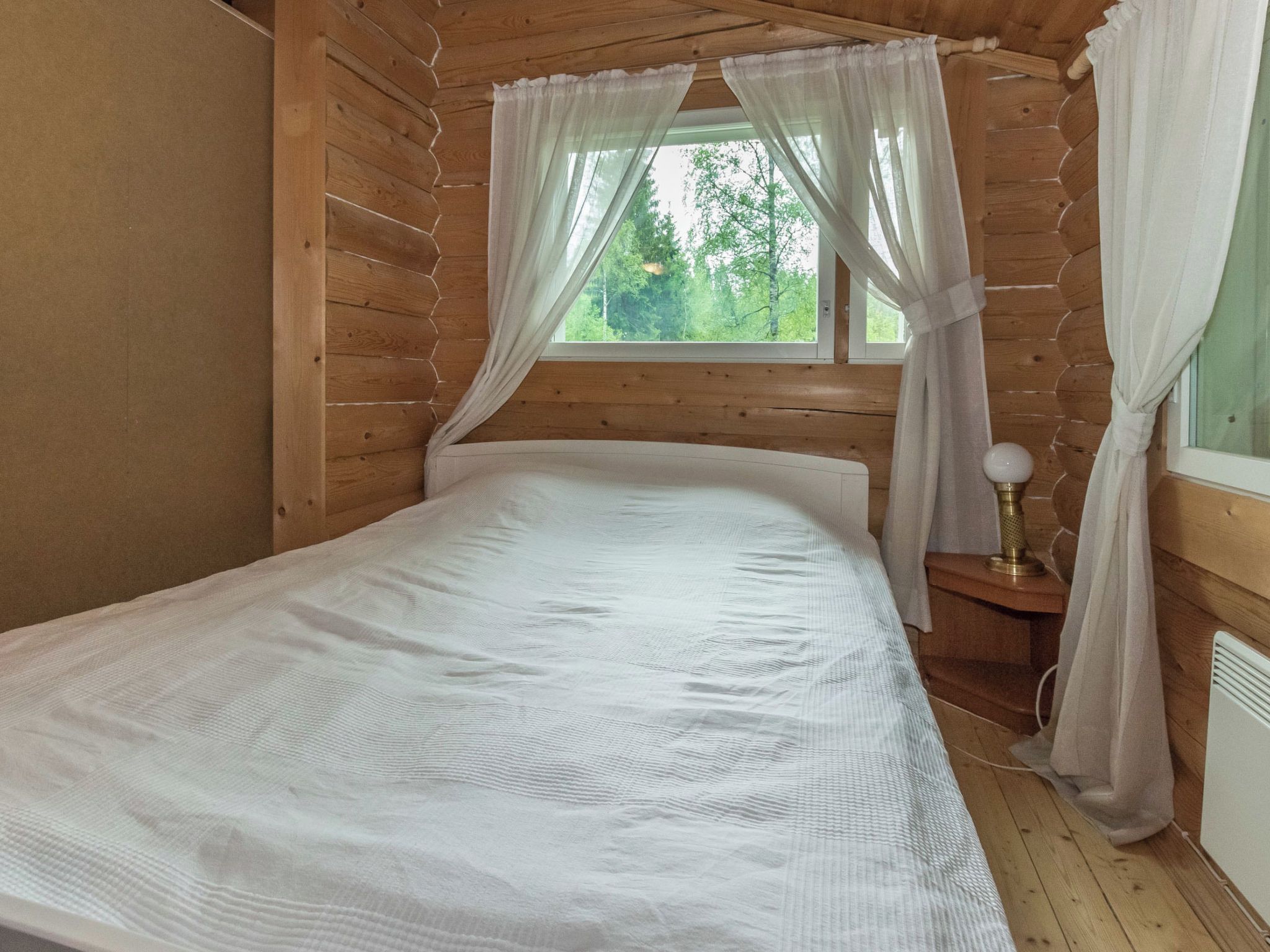 A wooden cabin room with a bed, a window with curtains, and a nightstand with a lamp.