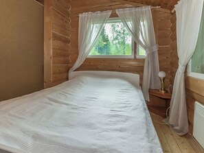A wooden cabin room with a bed, a window with curtains, and a nightstand with a lamp.