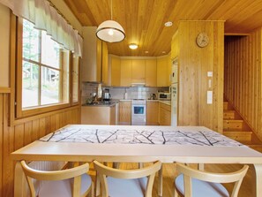 A wooden interior with a dining table, chairs, a kitchen with appliances, and a clock on the wall.
