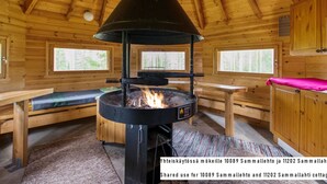 A wooden cabin interior with a central wood-burning stove, benches, and a window overlooking greenery.