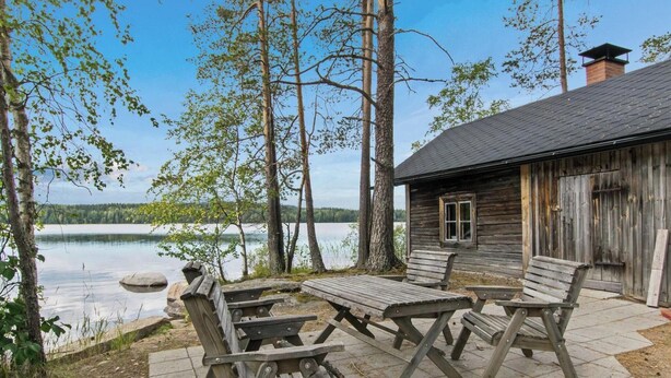 A rustic wooden cabin with a stone patio, surrounded by trees and overlooking a calm lake.