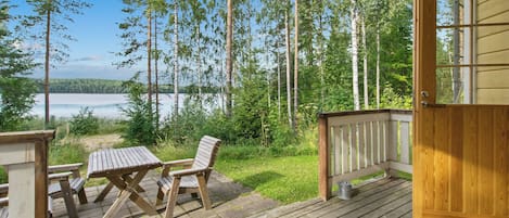 A wooden deck with a table and chairs, overlooking a lake with trees in the background.