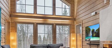 A spacious living room with a high ceiling, large windows, a fireplace, and a television.
