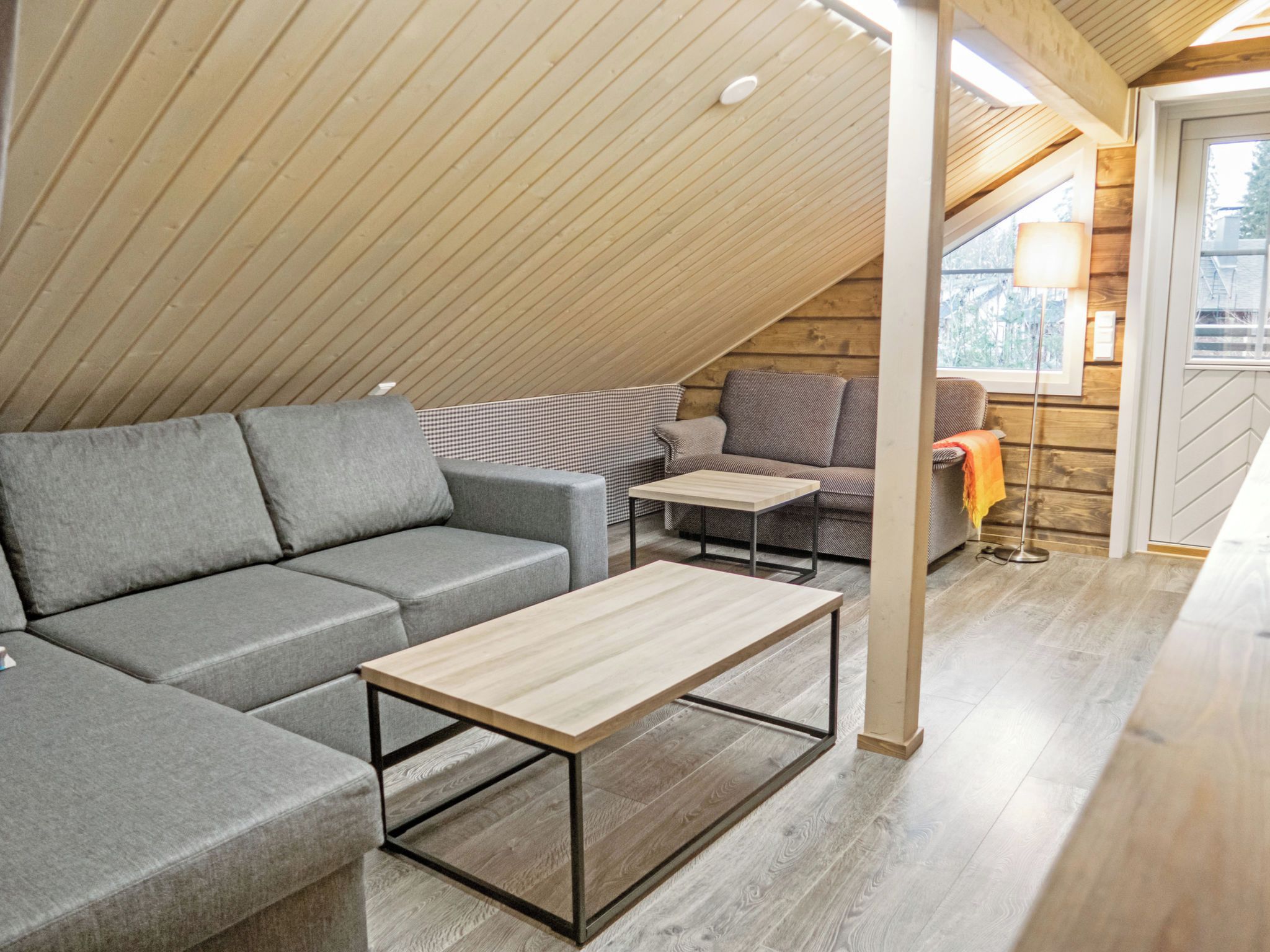 Un espai tipus loft amb sostre inclinat, un sofà modular gris, una taula de fusta petita i una finestra amb vista a l'exterior.