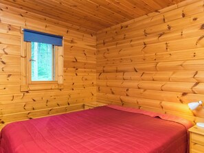 A wooden cabin room with a bed, a nightstand, a lamp, and a window with a blue blind. - Metsola / huilinpaikka by Interhome (Kerimäki)