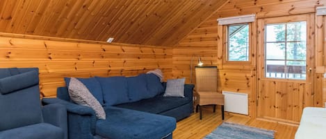 L’intérieur d’un chalet accueillant, avec un plafond en bois, un ensemble de canapés, un fauteuil et une fenêtre donnant sur les arbres.