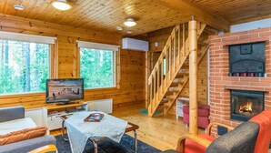 Interior de una cabaña con paredes de madera, chimenea, televisor y una escalera.