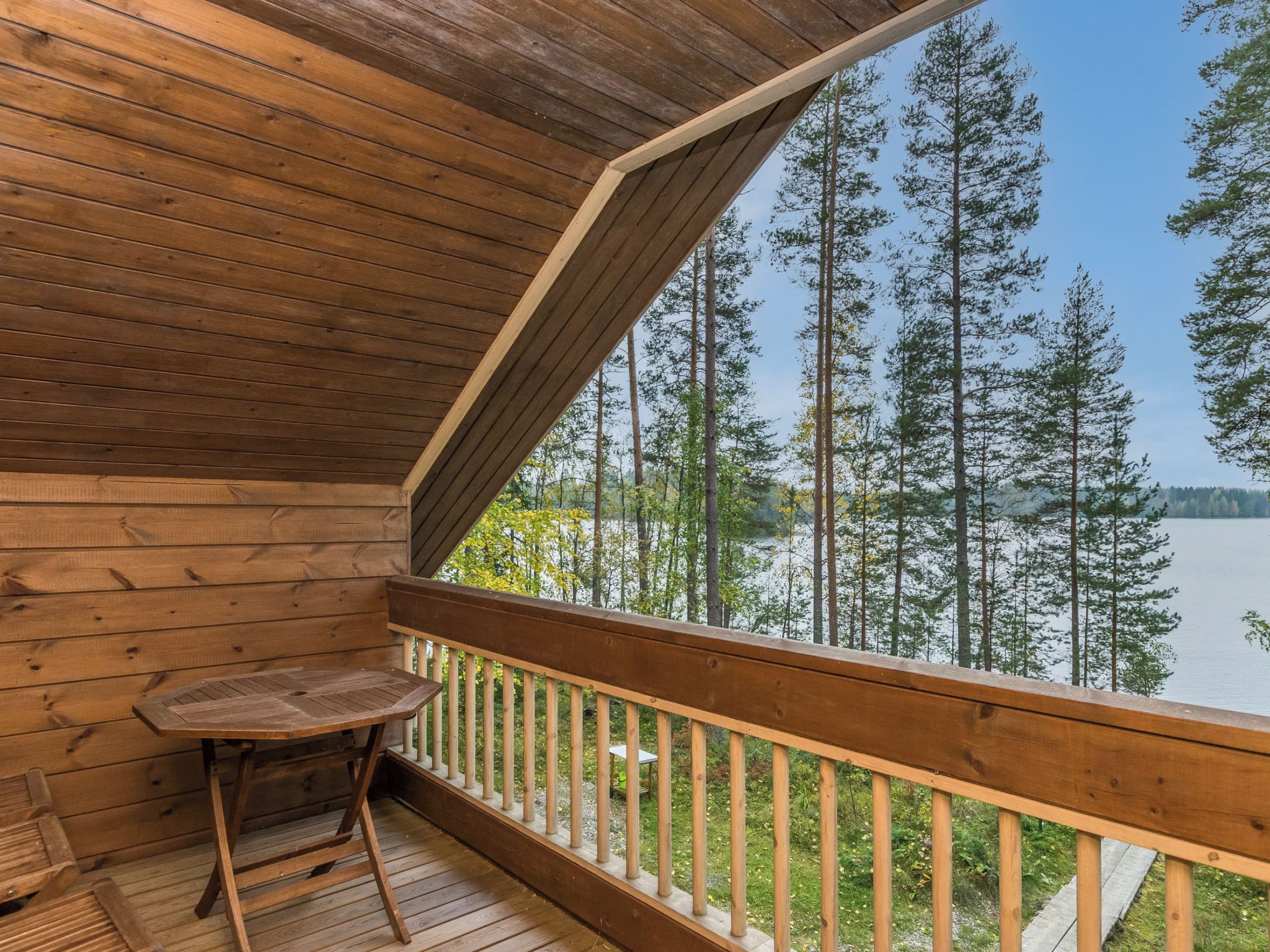 A wooden deck with a table and a chair, overlooking a forest and a lake.