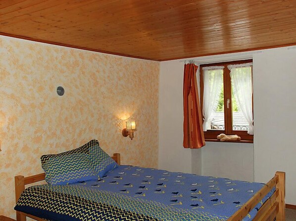 Une pièce comprenant un lit, une lampe fixée au mur, une fenêtre avec des rideaux et une vue sur la verdure à l’extérieur.