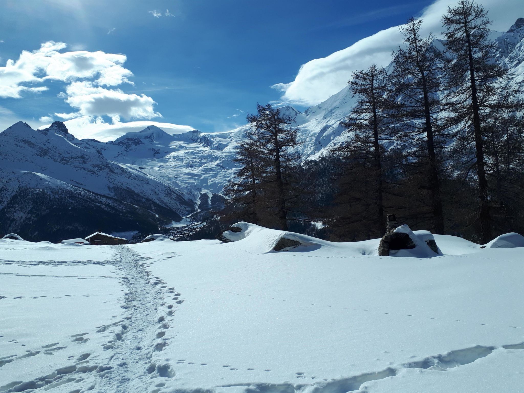 Un sentier de montagne enneigé, parsemé d’empreintes, bordé d’arbres et surplombant un ciel dégagé.