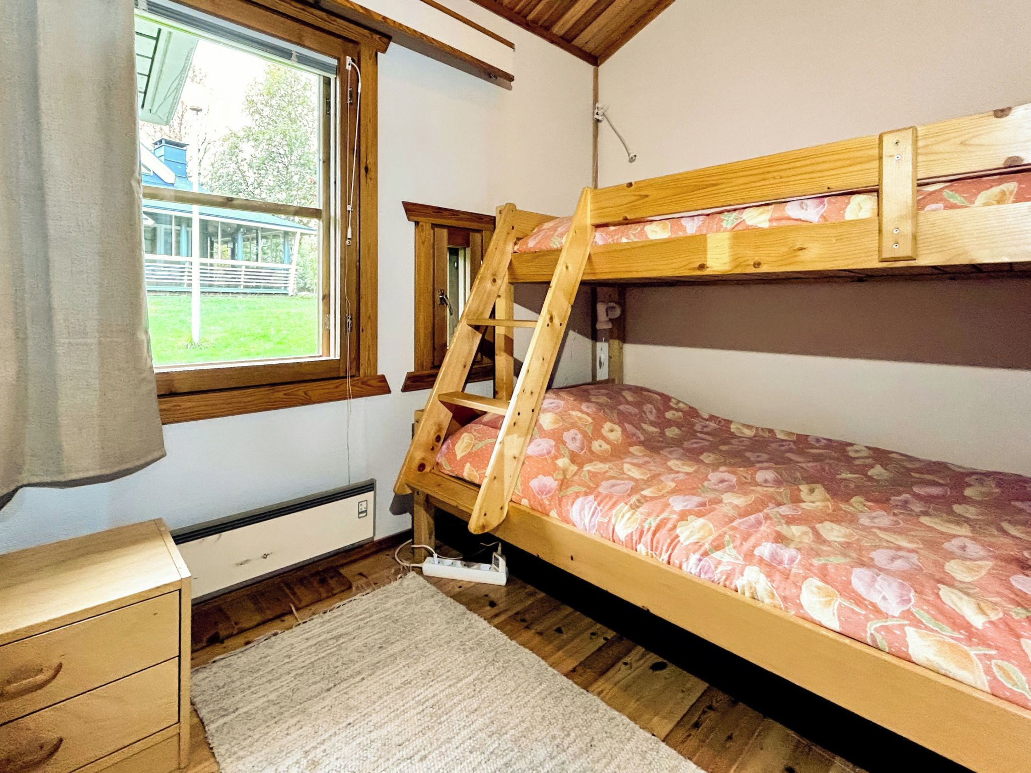 Uma cama beliche com colchão estampado com flores, uma escrivaninha de madeira, uma janela com vista para um edifício e vegetação, e um radiador sob a escrivaninha.