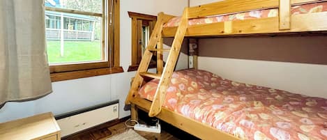 Una cama tipo litera con un colchón estampado con motivos florales, un escritorio de madera, una ventana con vistas a un edificio y zonas verdes, y un radiador debajo del escritorio.
