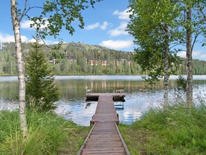 Un muelle de madera se adentra en un lago tranquilo, rodeado de abedules y con una zona residencial colinas al fondo.