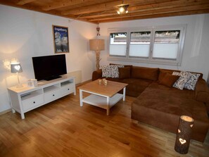 Een ingerichte woonkamer met een televisie, een wit kastje, een bruine bank, een salontafel en een raam met jaloezieën.
