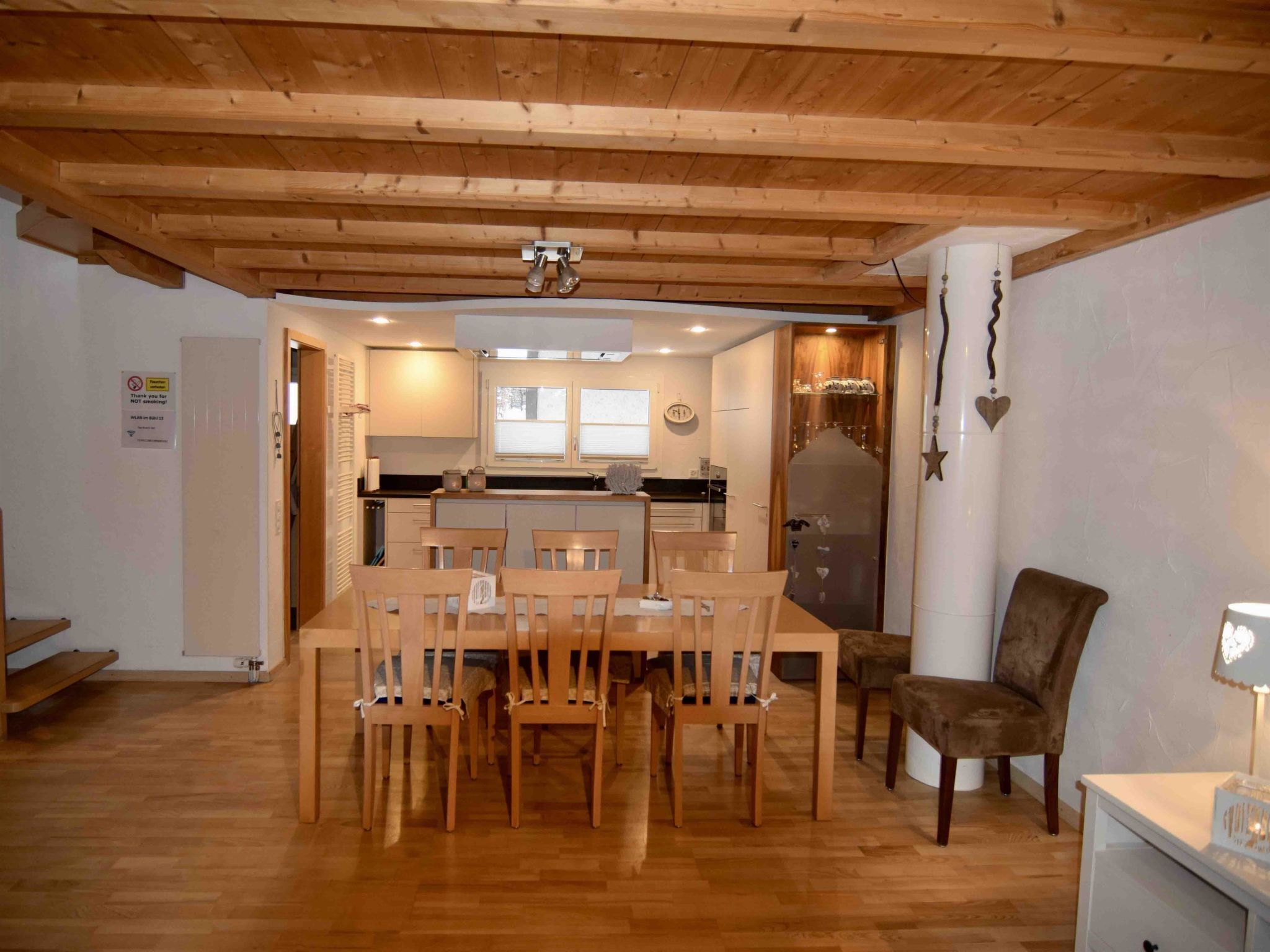 Uma área de refeições com uma mesa e cadeiras de madeira, uma cozinha ao fundo e uma parede branca com um aviso.