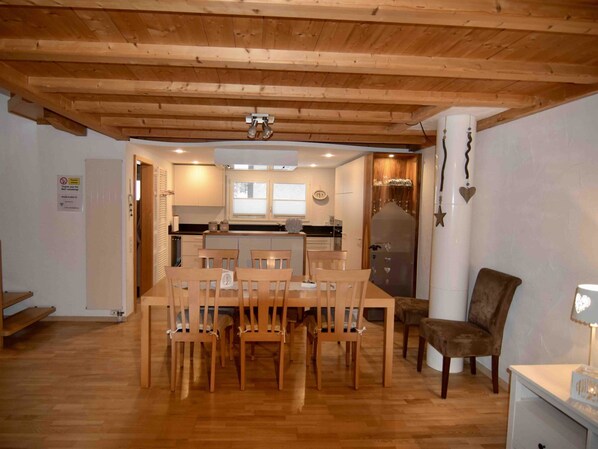 A dining area with a wooden table and chairs, a kitchen in the background, and a white wall with a sign.