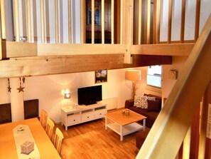A spacious living area with a wooden ceiling, a television on a white cabinet, a sofa with patterned cushions, and a dining table with chairs.