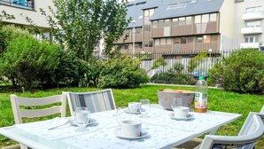An outdoor dining area with a table set for two, surrounded by greenery and modern buildings in the background.