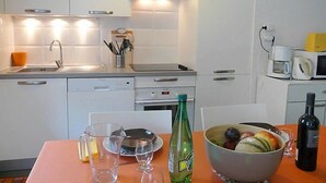A modern kitchen with white cabinets, a stainless steel sink, and a microwave. There is a dining table with a bowl of fruit and a bottle of water.