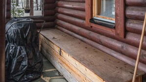 L’intérieur d’une cabane en rondins, avec un banc, un objet recouvert d’un tissu et une fenêtre avec des stores.