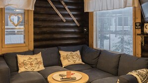 Un salon chaleureux avec une tête de lit en bois foncé, un canapé d’angle gris agrémenté de coussins à motifs et une petite table d’appoint en bois sur laquelle repose une télécommande.