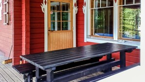 A wooden deck with a picnic table and benches, a red building with a wooden door, and a window with a view of greenery. - Vaahtera by Interhome (Kaavi)