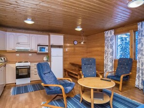 A compact kitchen with a stove, microwave, and refrigerator. There are two blue chairs around a small table, and a window with curtains. - Vaahtera by Interhome (Kaavi)