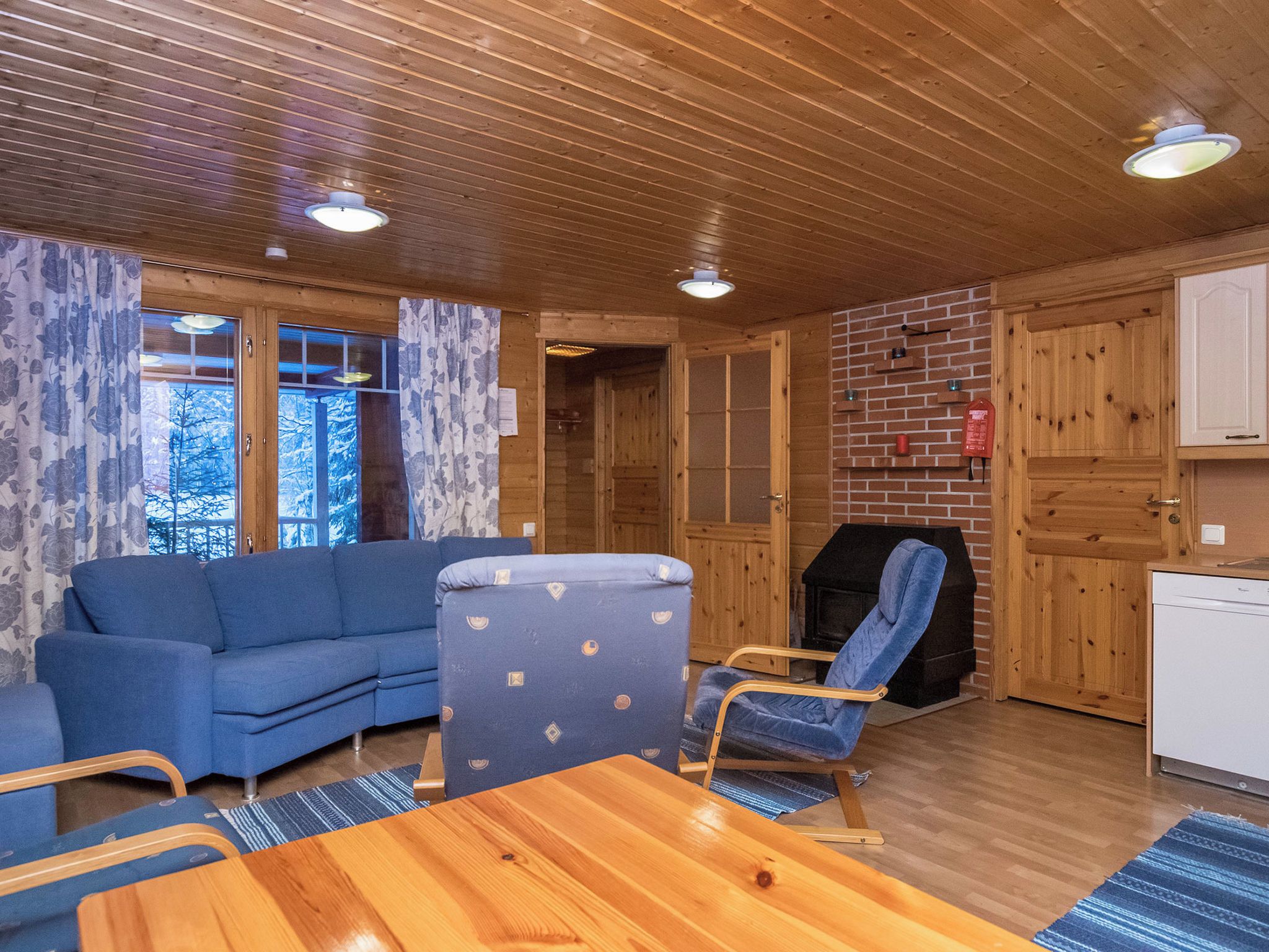 Ein gemütliches Blockhaus-Interieur mit Holzbalkendecke, einem blauen Sofa, einem blauen Sessel, einem Holzesstisch und einem Kamin aus Ziegelsteinen.