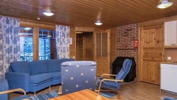 A cozy cabin interior with a wooden ceiling, a blue sofa, a blue armchair, a wooden dining table, and a brick fireplace.