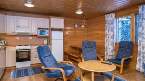 A compact kitchen with white cabinets, a microwave, a stove, and a refrigerator. There are two blue chairs and a small round table with a remote control. A clock is visible on the wall. - Vaahtera by Interhome (Kaavi)