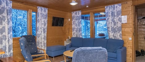 Ein gemütliches Blockhauskamin-Interieur mit Holzbalkendecke, einem blauen Sofa, einem blauen Sessel, einem kleinen Tisch und einem an der Wand befestigten Fernseher.