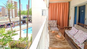 A balcony with a wooden table, a patterned sofa, and a view of a swimming pool and palm trees. - El Junco by Interhome (Benidorm)