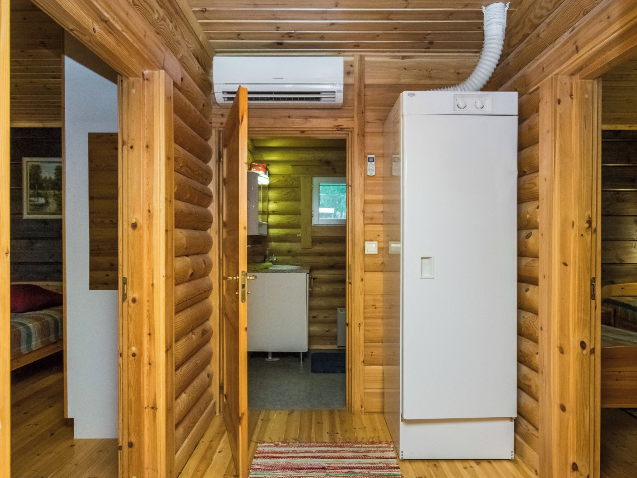 A log cabin interior with a wooden exterior, a central hallway leading to a kitchen area, and a white refrigerator.