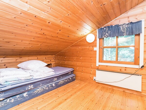 A small, cozy room with wooden walls and a sloped ceiling, featuring a single bed with pillows and a mattress, a window with a view of greenery, and a wall-mounted heater. - Ruka-linkki 3a by Interhome (Kuusamo)